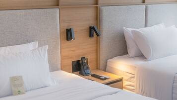 Una habitación de hotel con dos camas, cabecero de madera, mesita de noche con teléfono y control remoto, y una lámpara de pared.