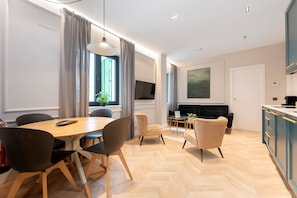 A modern living room with a dining area, a flat-screen TV mounted on the wall, and a kitchen with blue cabinets.