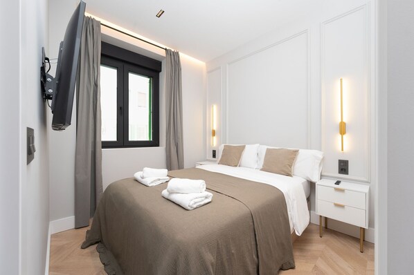 A hotel room with a bed, bedside tables, a window with curtains, and wall-mounted lighting.