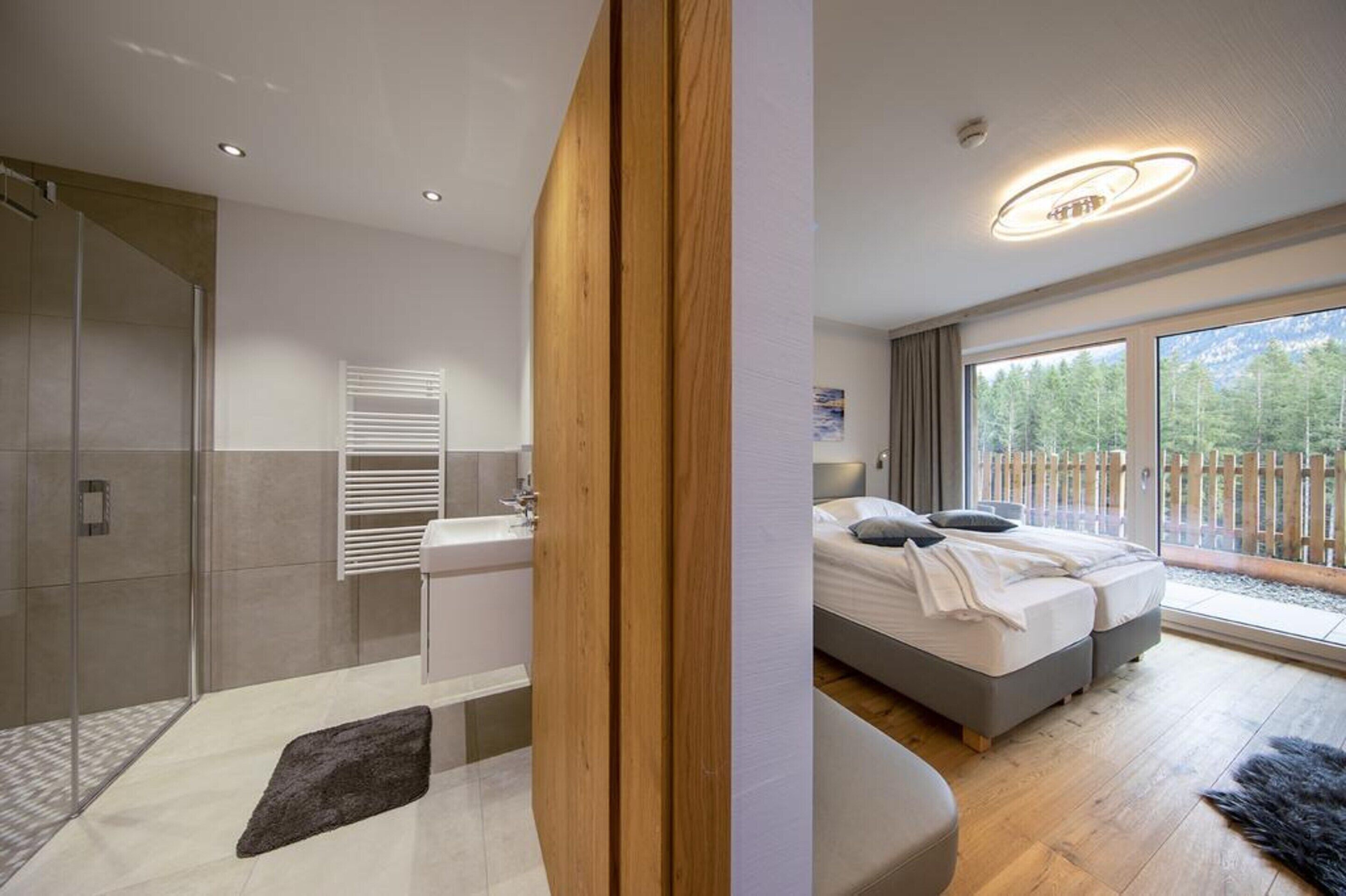 A modern bedroom with a large bed, a bathroom with a glass shower enclosure, and a view of a wooden deck.