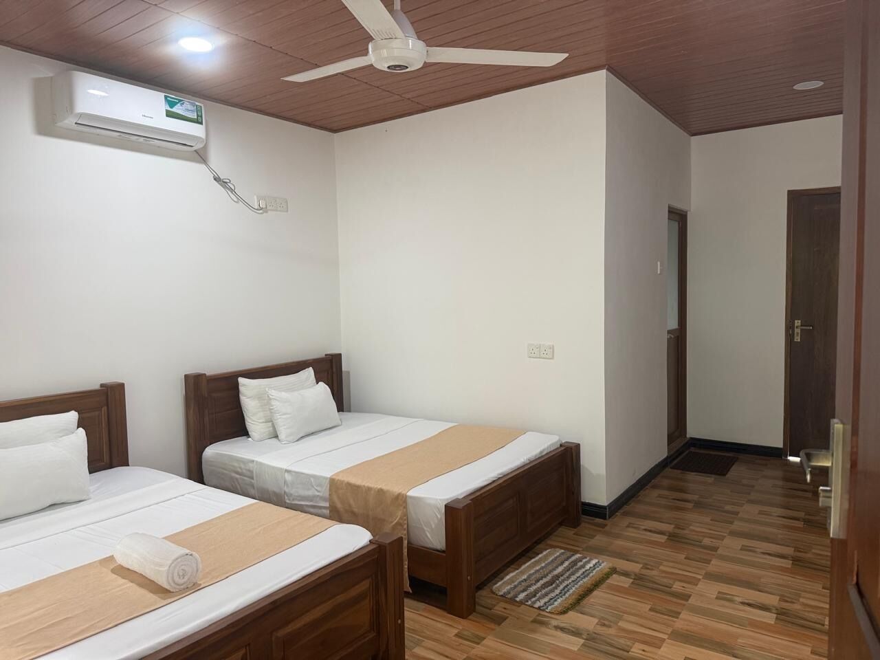 Ein Hotelzimmer mit zwei Einzelbetten, einem Deckenventilator, einer Klimaanlage und einem Holzboden.