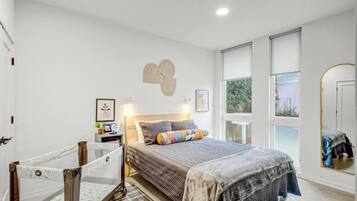 A bedroom with a bed, a crib, a nightstand, and a window with a view of greenery.