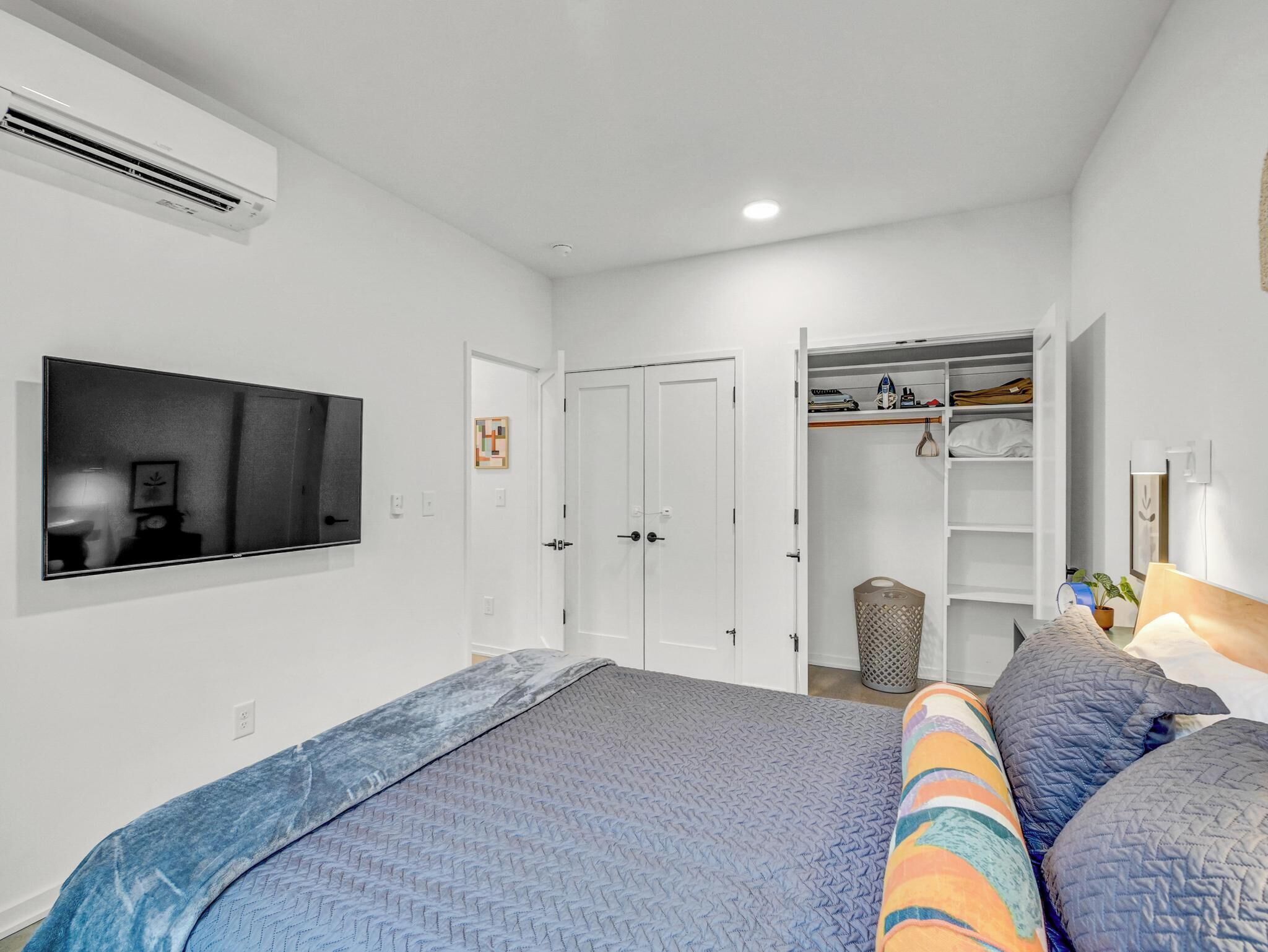 Un dormitorio moderno con una cama grande, un televisor instalado en la pared, un armario con estantes y un pequeño cubo de basura.