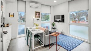 A modern, well-lit room with a dining area, a sofa, a TV, and large windows offering a view of the outside. - Mini Metro - Downtown Rooftop with Skyline View! (Raleigh)