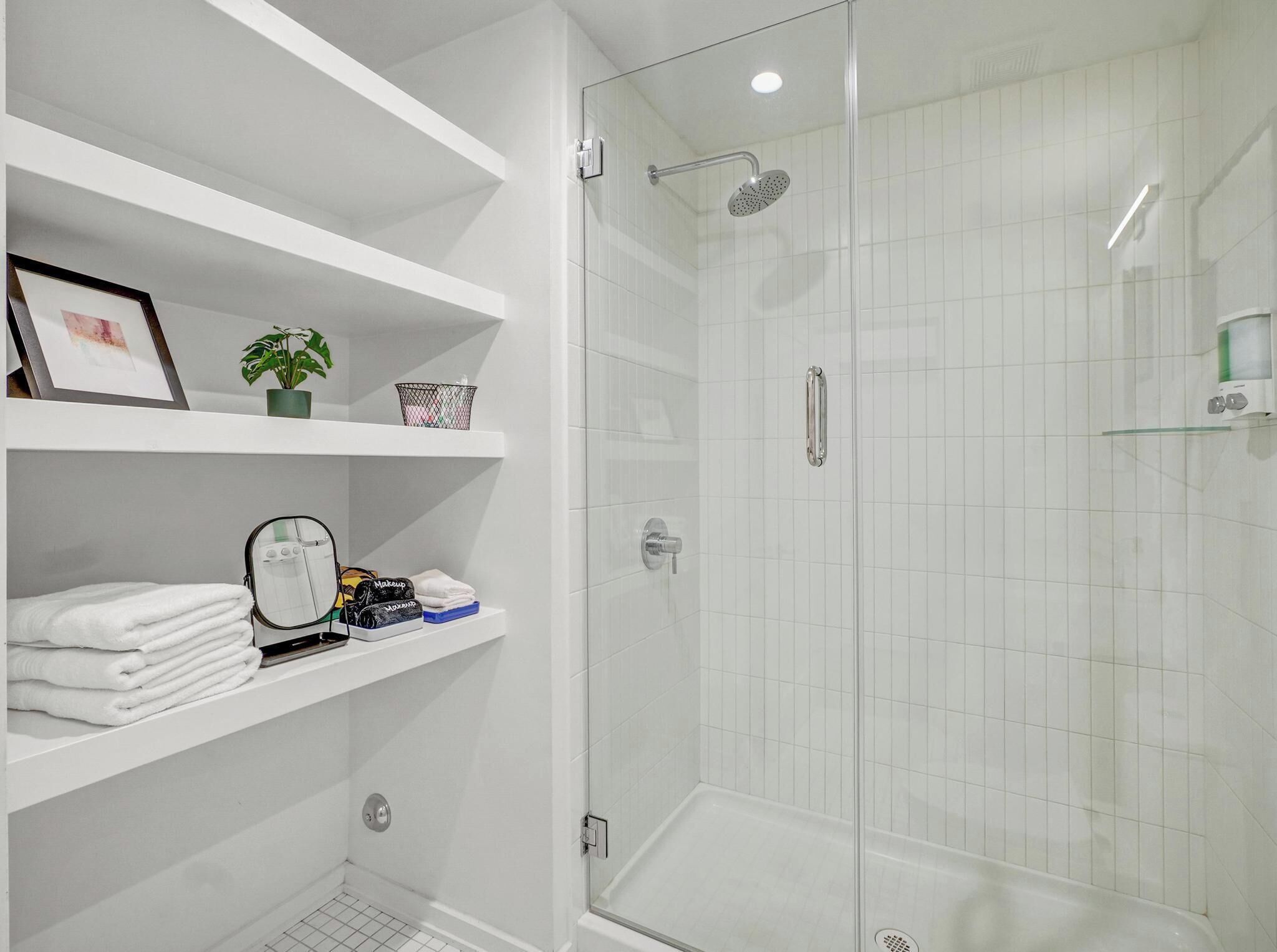 Ein modernes Badezimmer mit einer in Glas eingefassten Dusche, weißen Fliesen und Regalen mit Handtüchern und Pflegeprodukten.