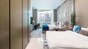 A modern hotel room with a large bed, a desk with a chair, a sofa, and a view of the cityscape through large windows. - Luxury Canal-View Studio (Business Bay)