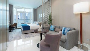 A modern hotel room with a bed, a grey sofa, a round coffee table, and a chair. There is a large window with a view of buildings. - Luxury Canal-View Studio (Business Bay)
