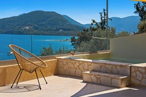 A balcony with a wicker chair, a glass table, and a view of a lake and mountains.