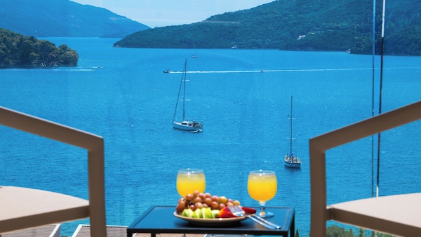 A table with a fruit plate and two glasses of orange juice, overlooking a calm sea with boats and green hills in the background. - MANDRAGORAS ECO FRIENDLY (Perigiali)