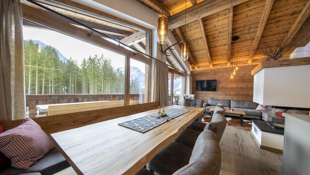 A modern interior with a wooden ceiling, large windows offering a mountain view, a wooden dining table, and a sofa. - Chalet in Neukirchen Near Ski Lift (Neukirchen am Grossvenediger)