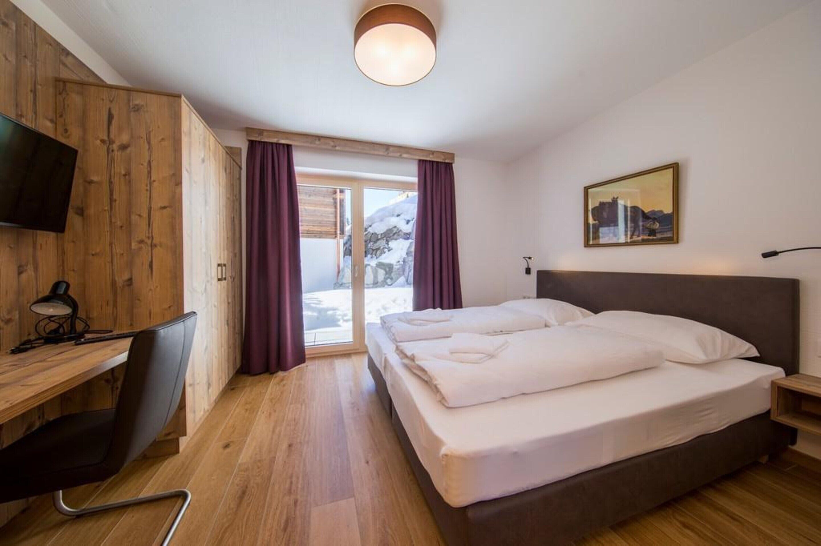 A hotel room with a wooden desk, a bed, a TV, and a window with curtains.