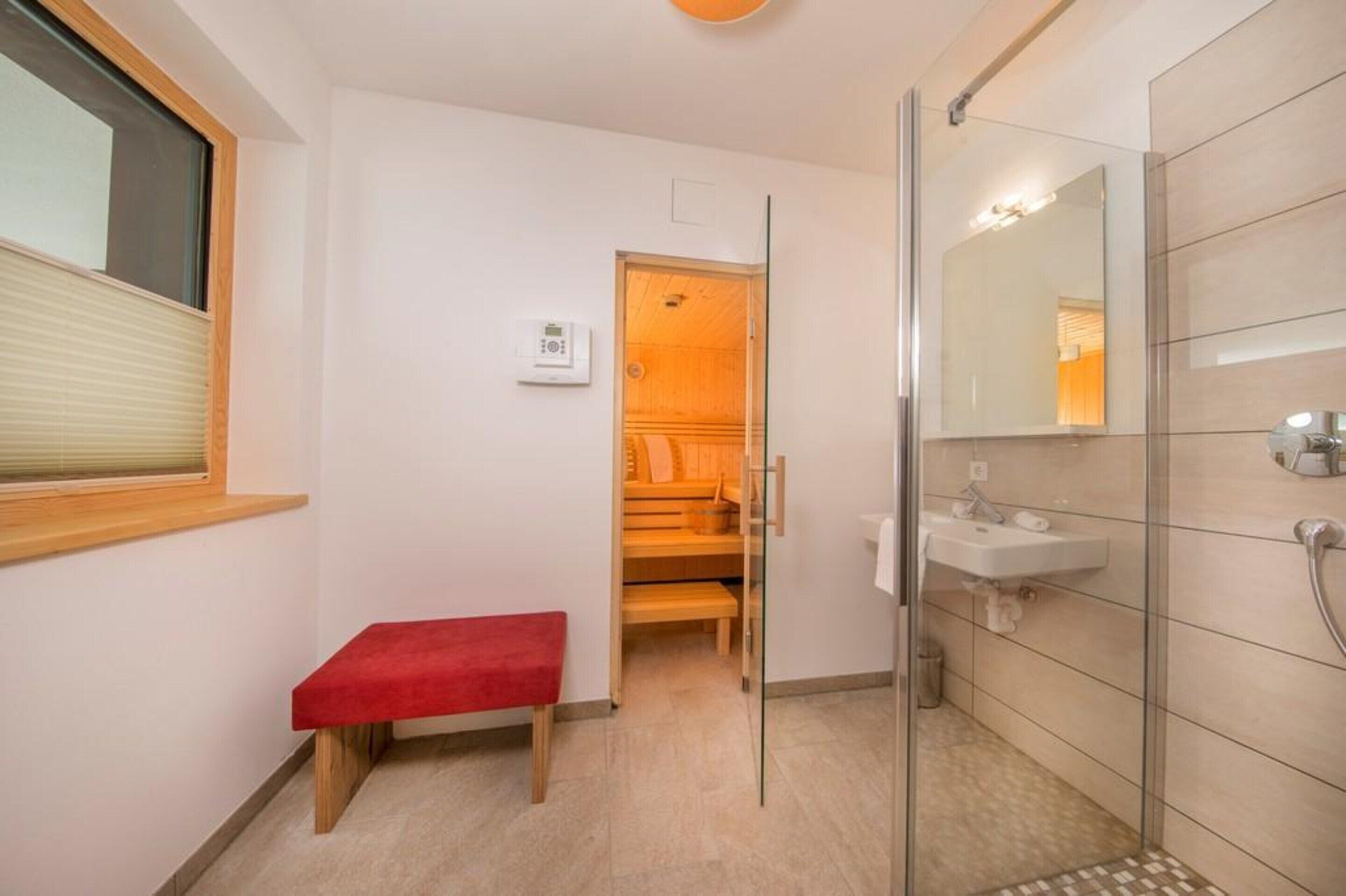 Un baño moderno con cabina de ducha de vidrio, un banco rojo, un banco de madera, un lavamanos blanco y un espejo.