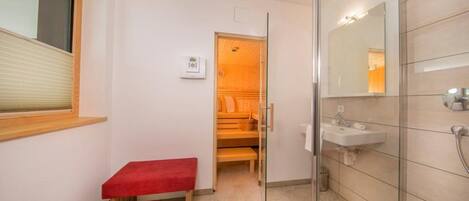 Un baño moderno con cabina de ducha de vidrio, un banco rojo, un banco de madera, un lavamanos blanco y un espejo.