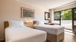 Habitación de hotel con dos camas, un escritorio con lámpara, un cuadro en la pared y un ventanal con vistas a árboles.