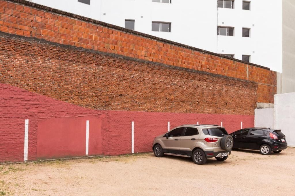 Un estacionamiento con dos coches aparcados junto a una pared roja y un edificio blanco al fondo.