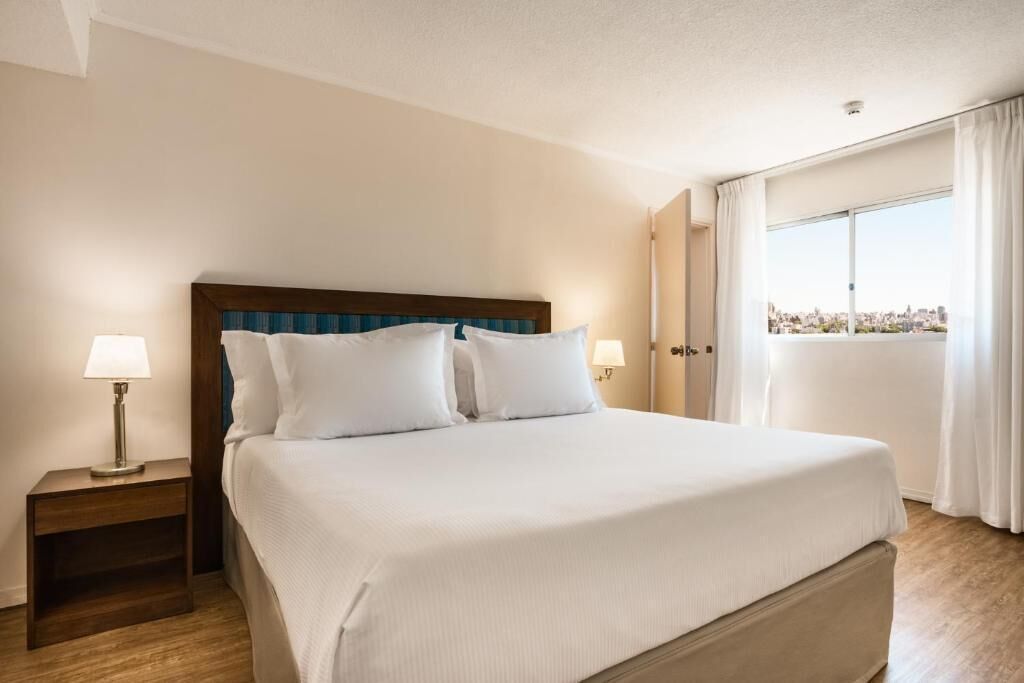 Habitación de hotel con una cama grande, dos lámparas de noche, un cabecero de madera y vistas a la ciudad a través de un ventanal amplio.