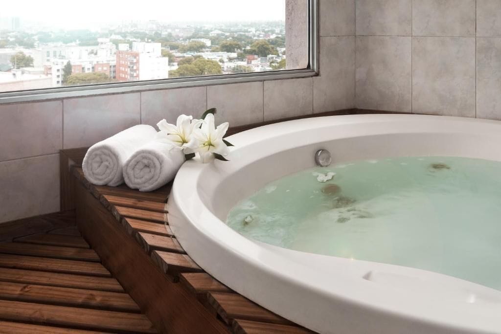 Un cuarto de spa con una bañera grande, escalones de madera y vistas de la ciudad a través de un ventanal.