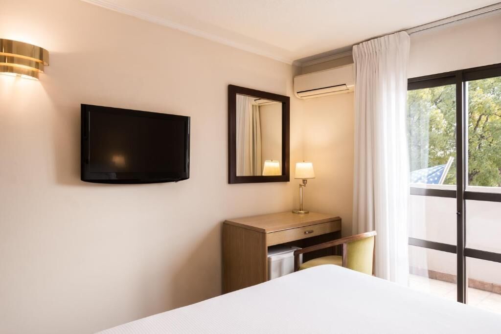 A hotel room with a bed, a TV mounted on the wall, a desk with a lamp, and a large window with curtains.