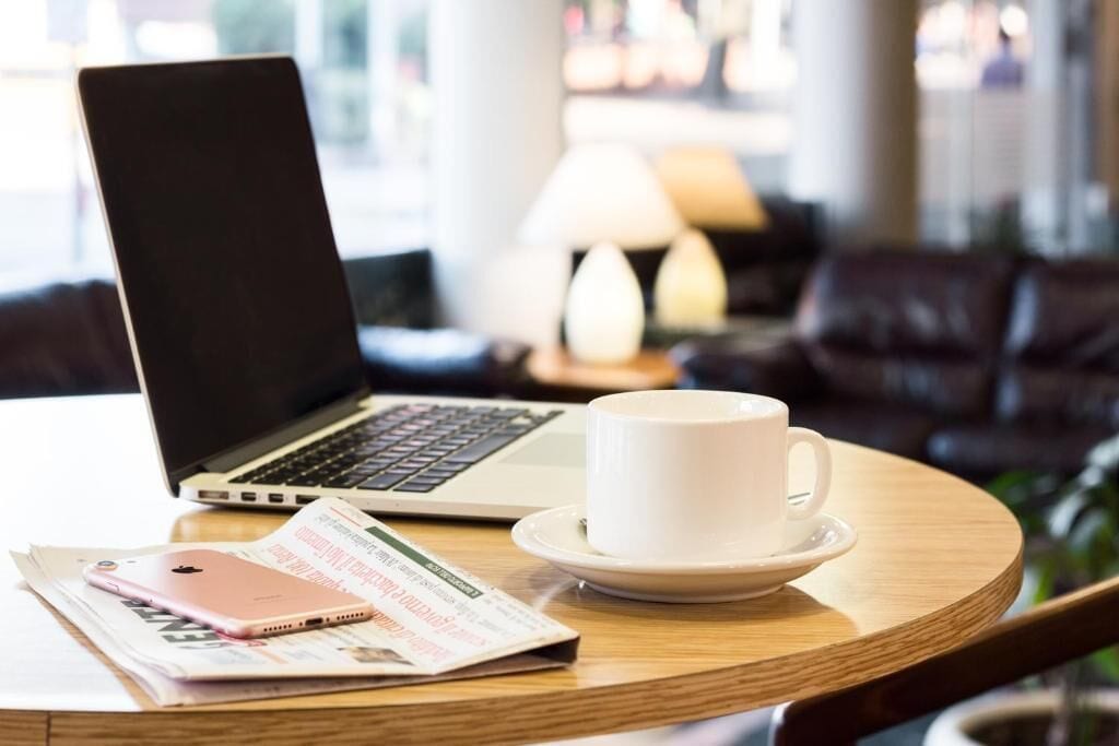 Una laptop sobre una mesa redonda de madera, con una taza y platillo blancos, un teléfono inteligente y una revista.