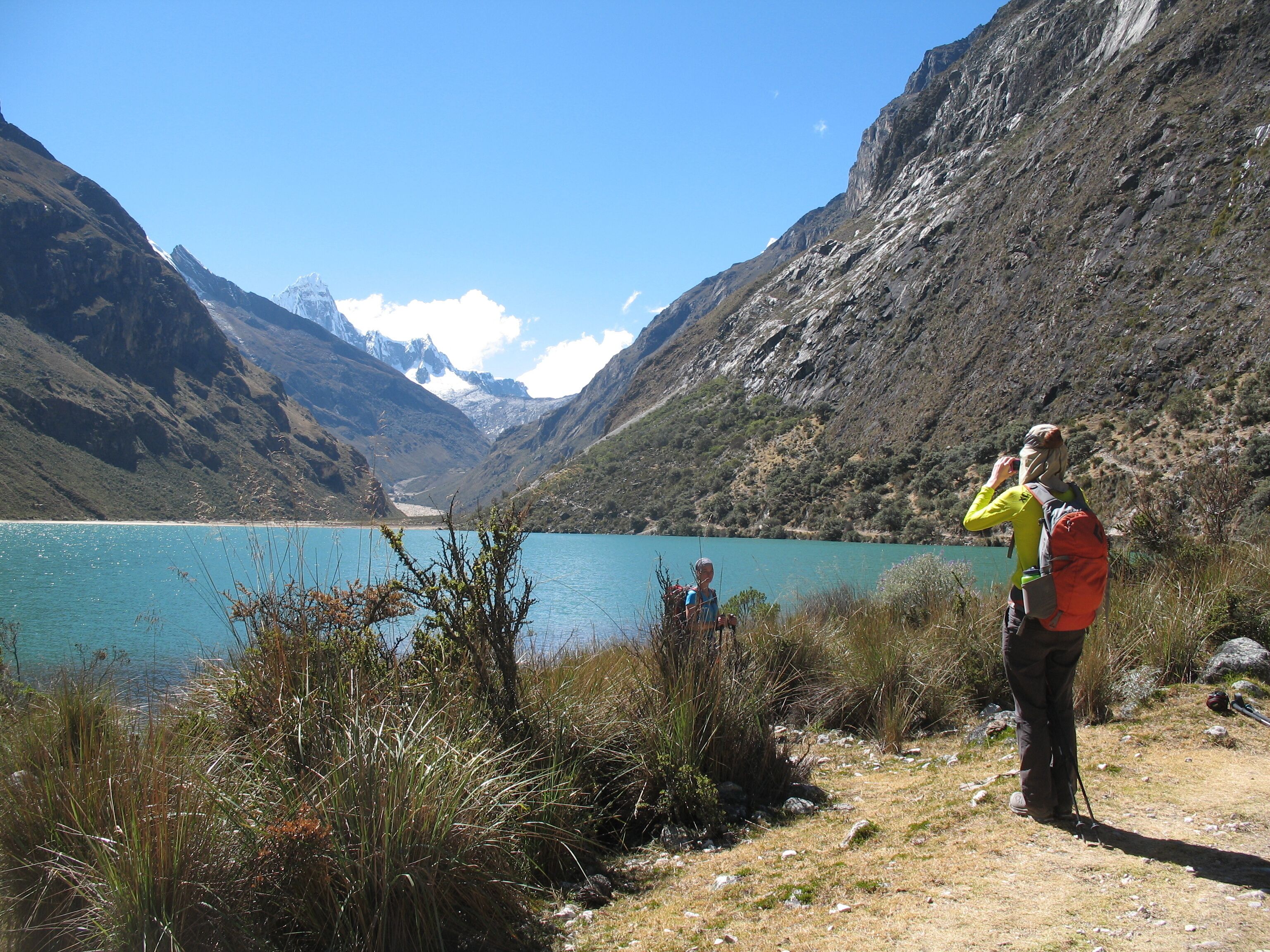 Un excursionista con chaqueta amarilla y mochila roja, de pie en una colina cubierta de hierba, con vistas a un lago turquesa y montañas al fondo.