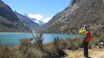 Un excursionista con chaqueta amarilla y mochila roja, de pie en una colina cubierta de hierba, con vistas a un lago turquesa y montañas al fondo.