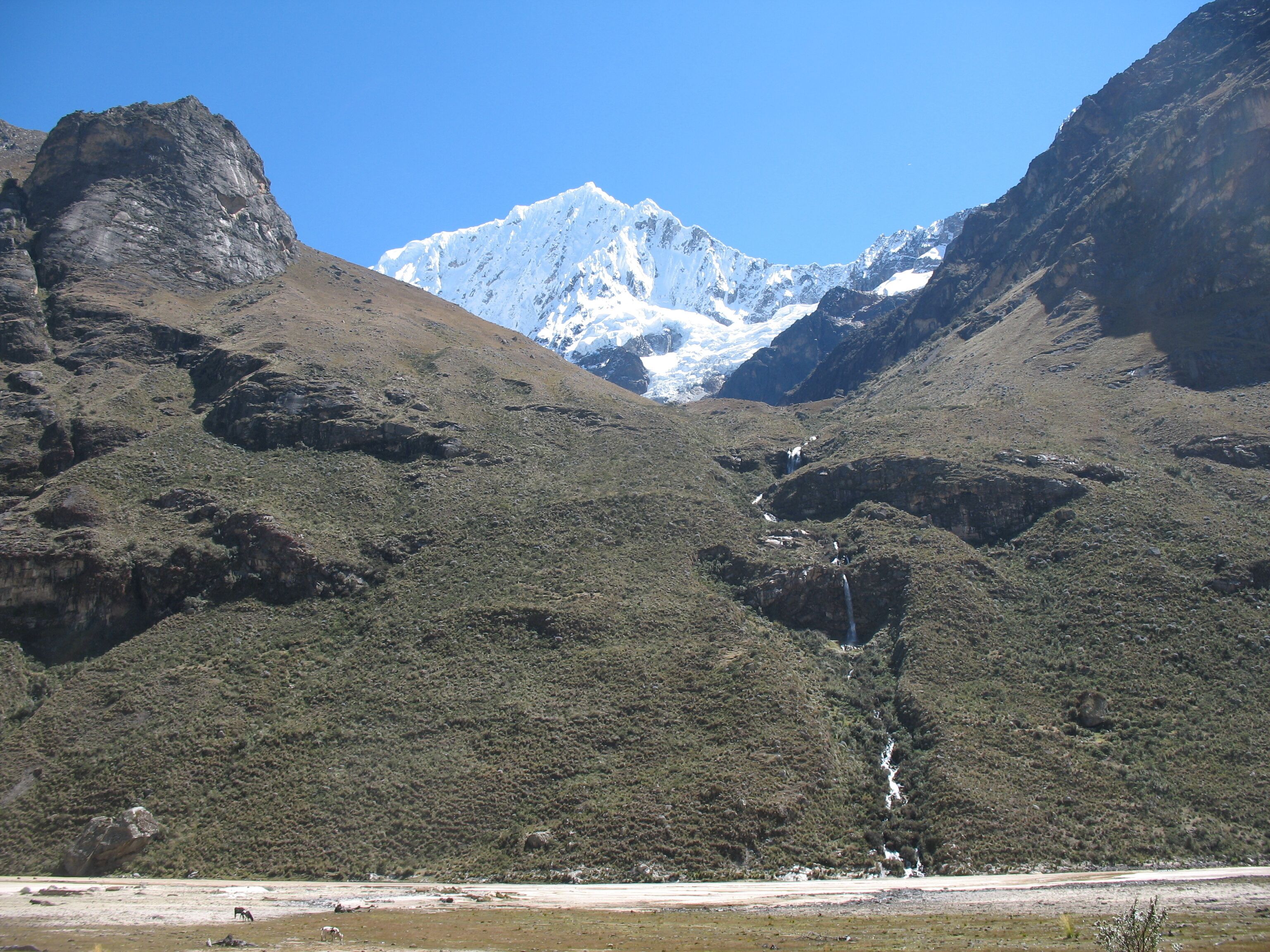 Eine gebirgige Landschaft mit einem markanten, schneebedeckten Gipfel im Hintergrund, felsigem Gelände und einem kleinen Wasserfall.