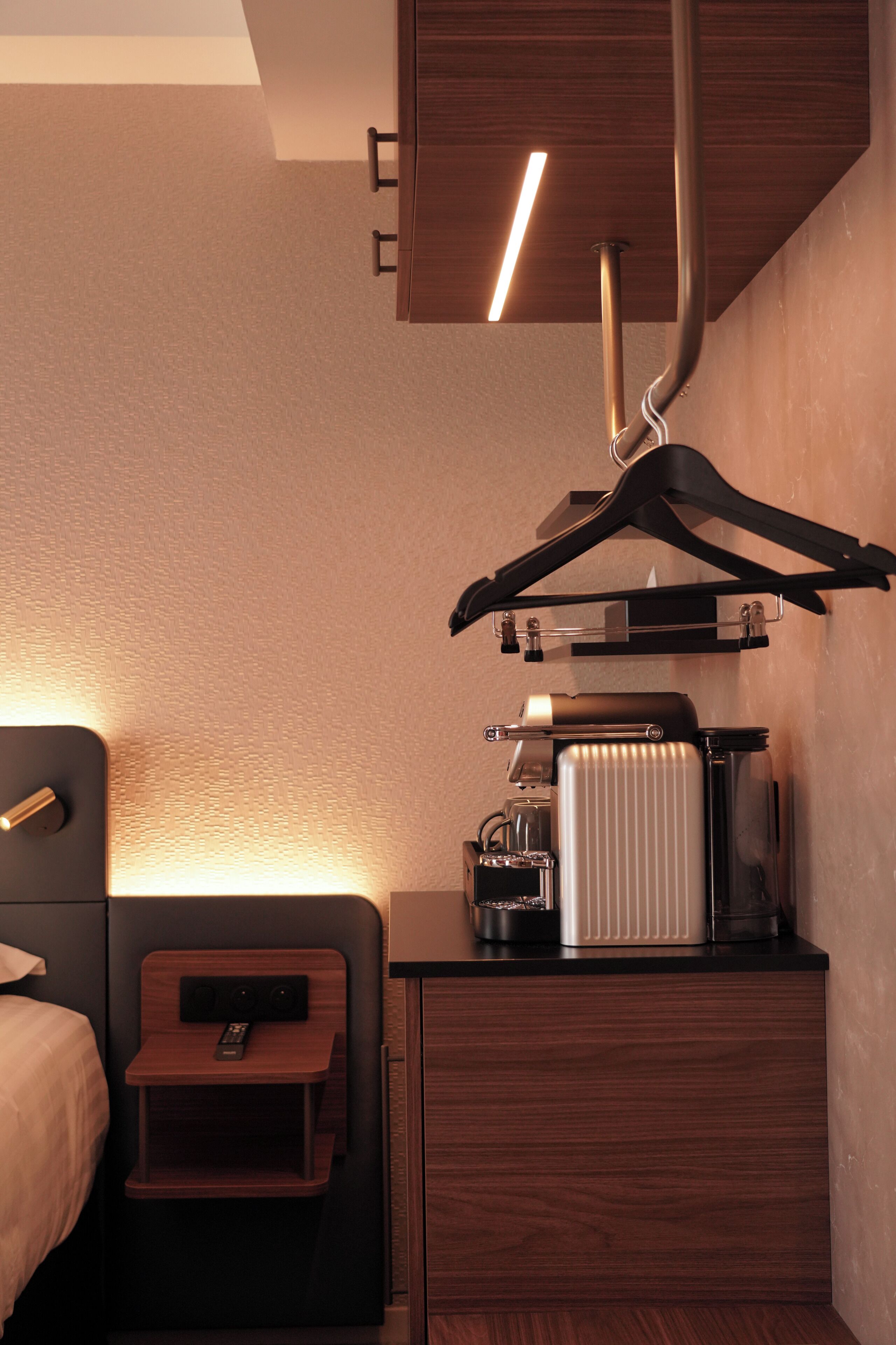 Una habitación de hotel con cama, mesita de noche y un mueble pequeño con hervidor y cafetera.