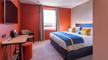 Quarto de hotel com cama, escrivaninha, cadeira, televisão e janela com cortinas.