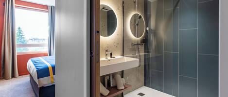 A modern bathroom with a glass-enclosed shower, a round mirror above the sink, and a bed with a blue and white striped bedspread.
