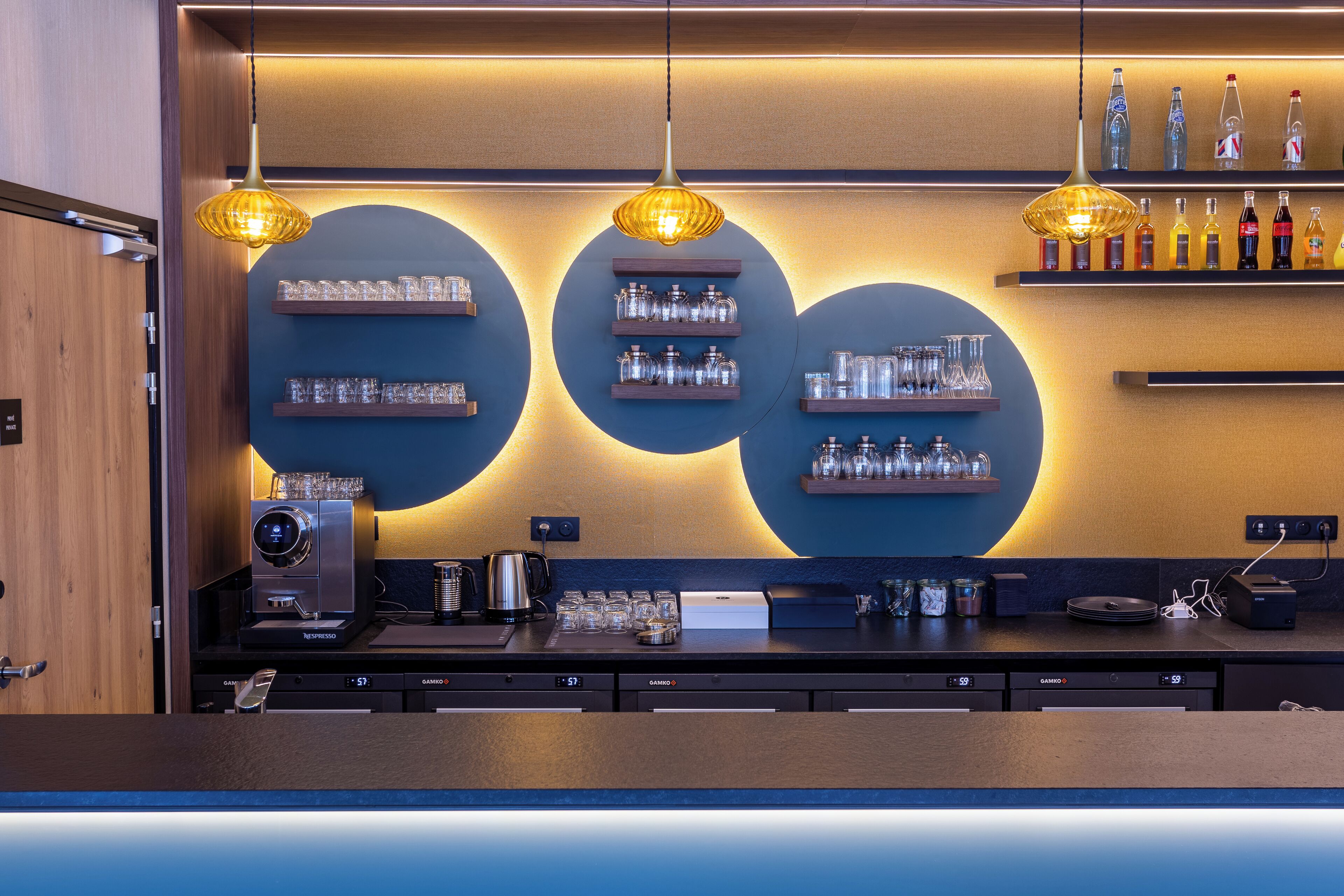 Zona bar moderna con macchina da caffè, scaffali con bicchieri e lampade a sospensione.