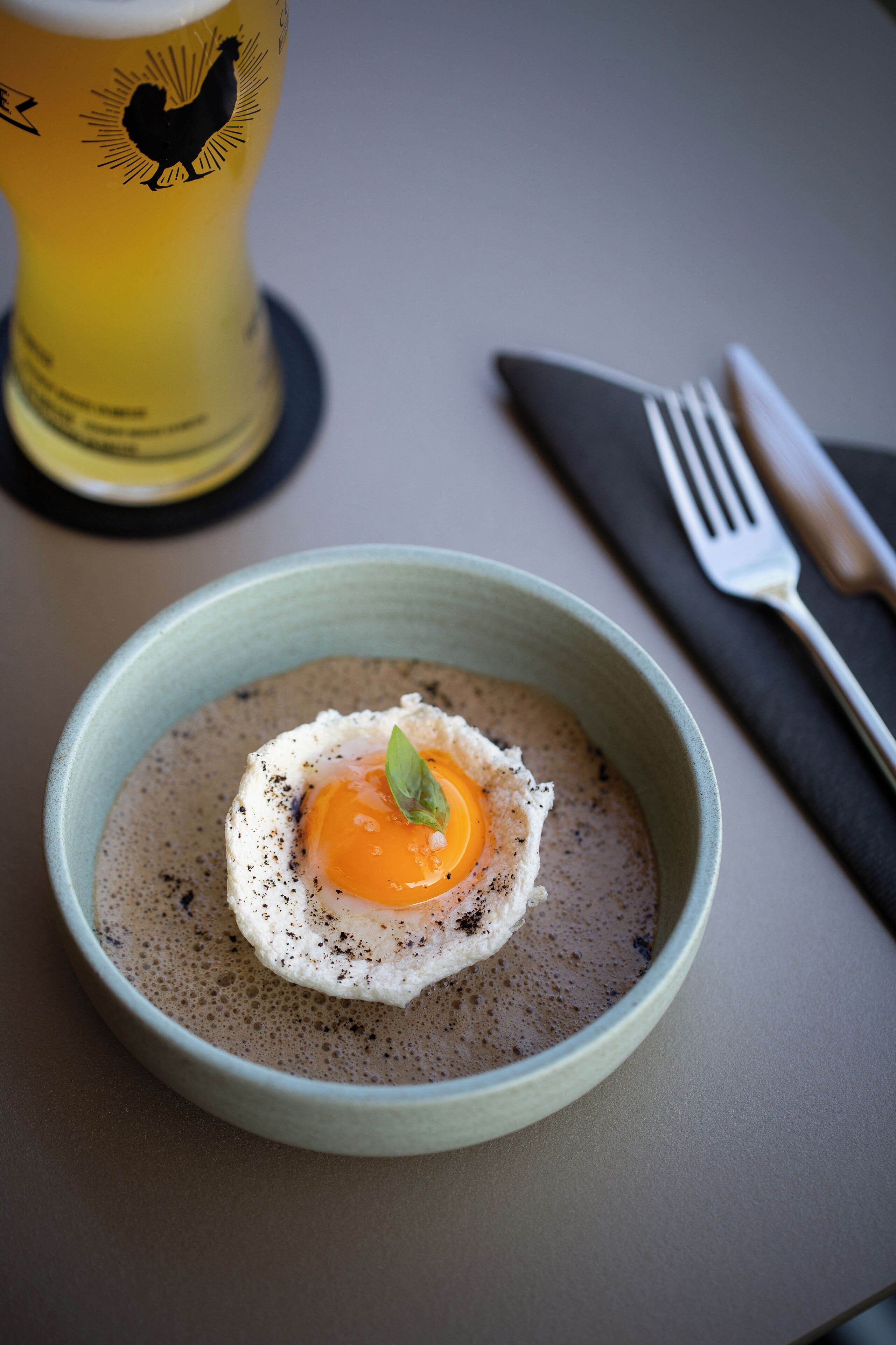 Un bol de nourriture surmonté d’un œuf frit, posé sur un lit de céréales, un verre de bière et des couverts sur une table.