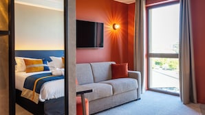 Una habitación de hotel con cama, sofá, televisión instalada en la pared y un ventanal con cortinas.