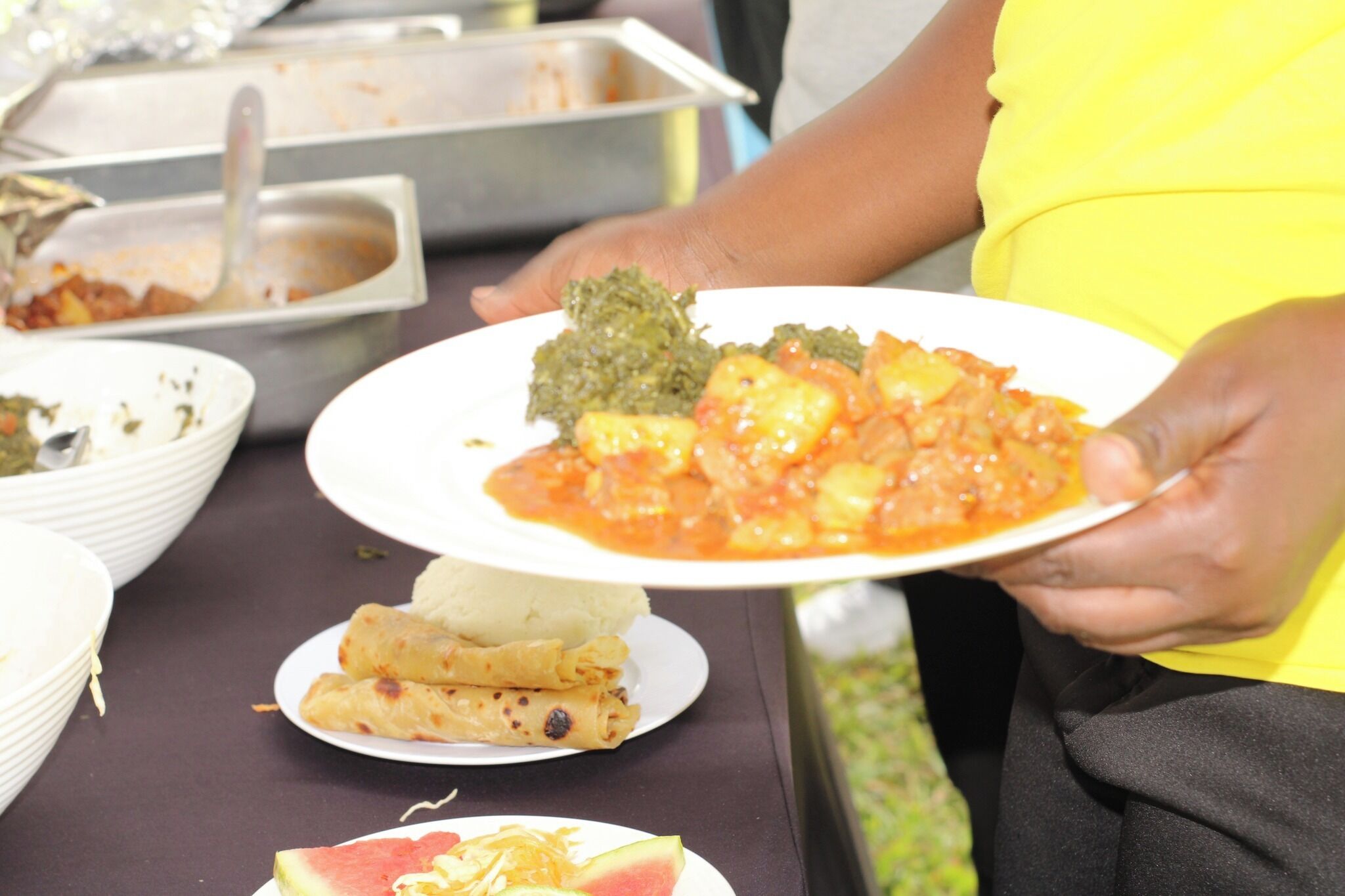 A person holding a plate with food, including a green dish and a red sauce with chunks, and a plate with rolled items and a slice of watermelon.