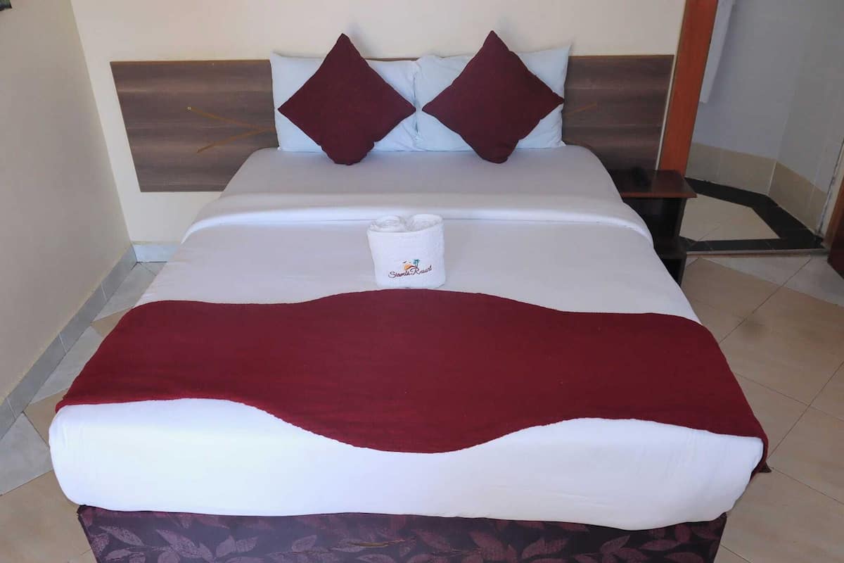 A neatly made bed with a white and red blanket, two dark red pillows, and a rolled white towel on the bed.