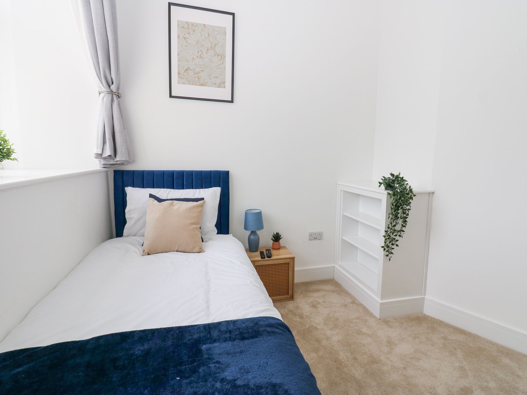 A bedroom with a bed, a bedside table, a lamp, a plant, and a wall-mounted shelf.