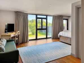 A modern hotel room with a large sliding glass door to the outdoors, a bed, a desk with a TV, and a sofa.