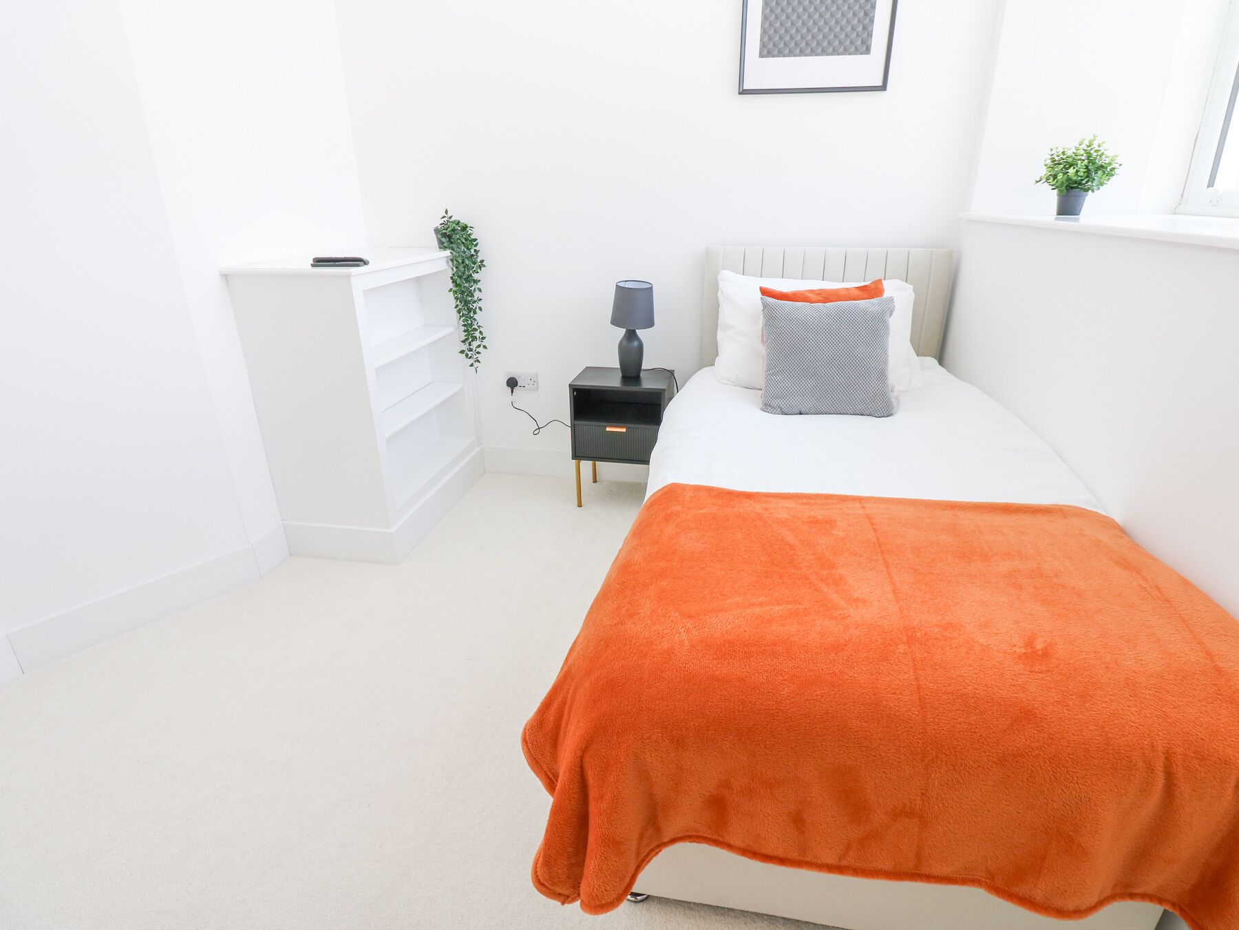 Un dormitorio con una cama, una mesita de noche, un estante blanco, una lámpara y una planta pequeña.