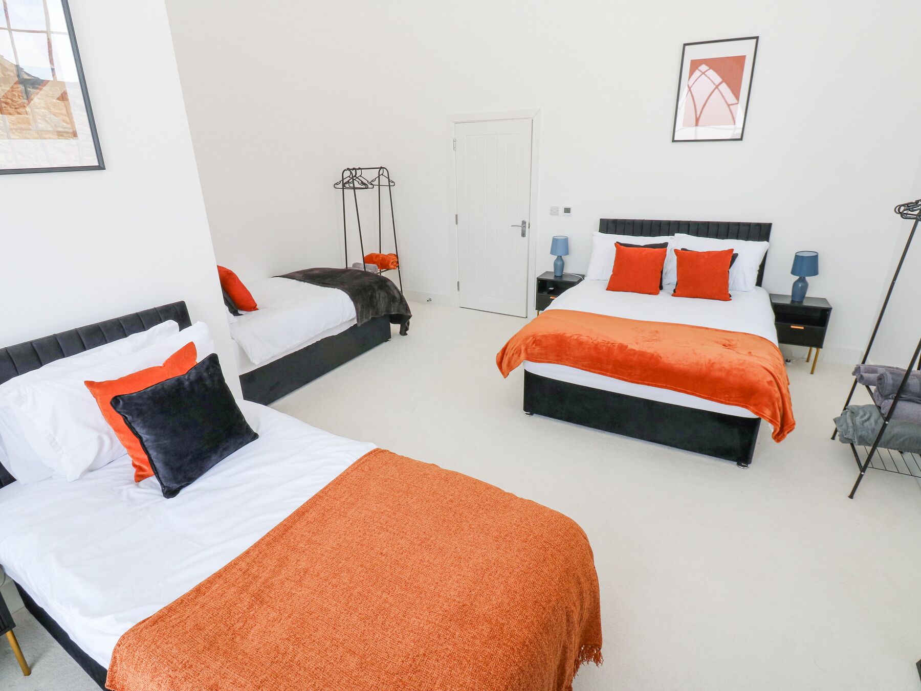A hotel room with two beds, each with white and orange bedding, a black headboard, and a small bedside table with a lamp.