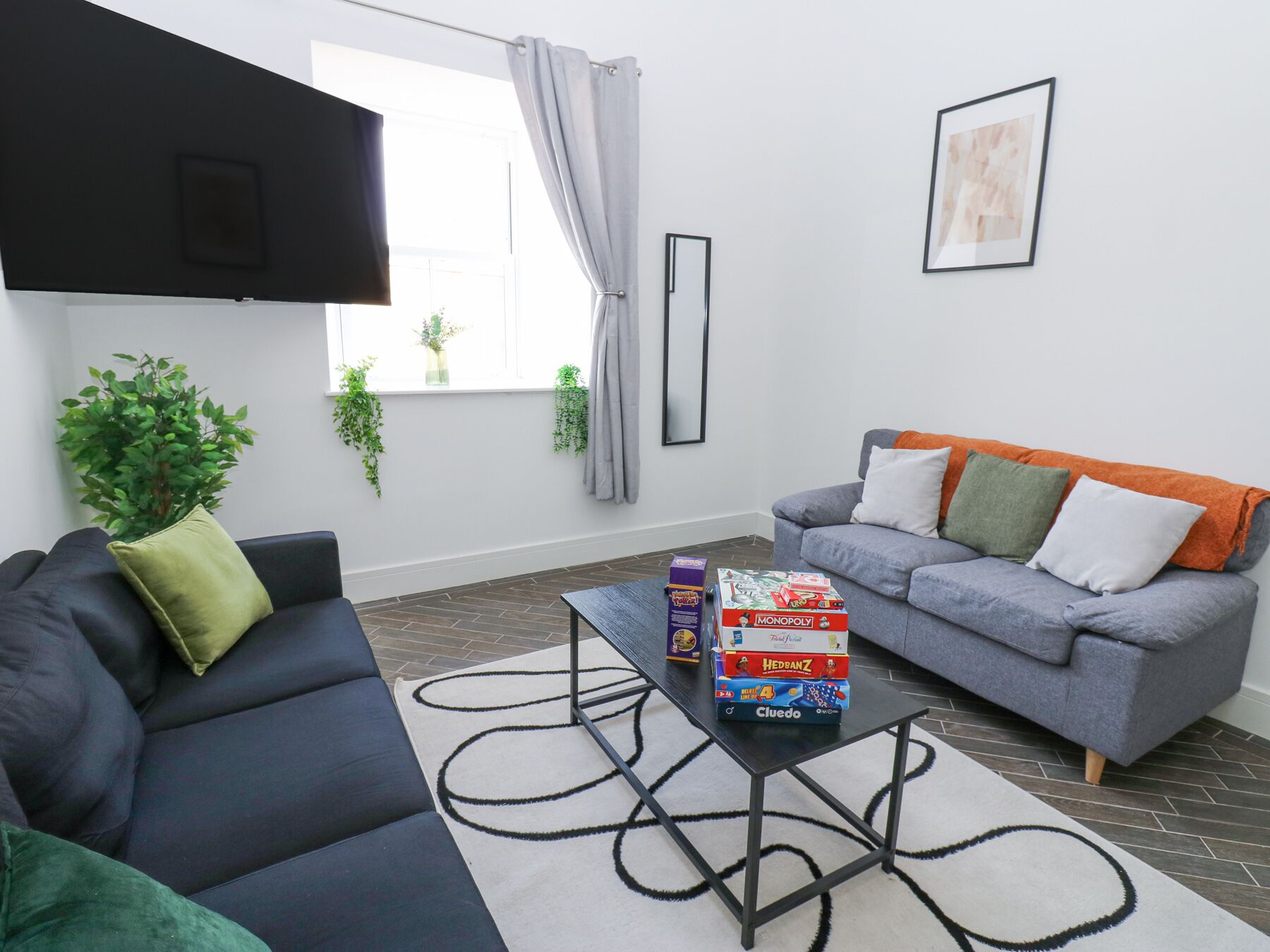 A modern living room with a dark sofa, a grey sofa with orange throw pillows, a coffee table with board games, and a large window with plants.