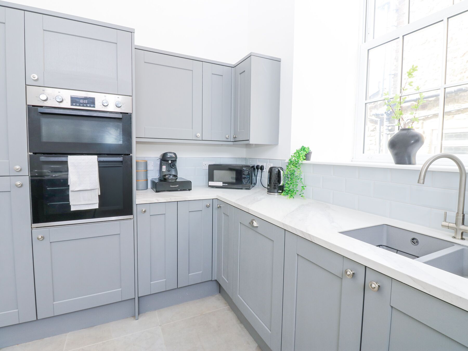 A modern kitchen with grey cabinets, a built-in oven, a coffee maker, a microwave, and a sink.