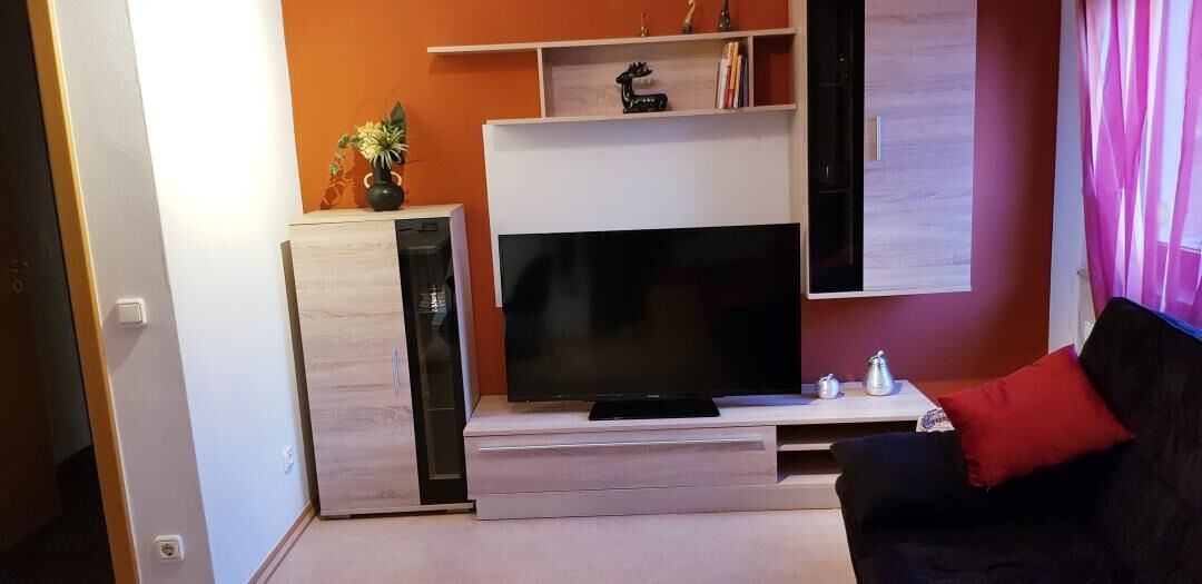 A living room with a flat-screen TV, a black sofa, and a red cushion.