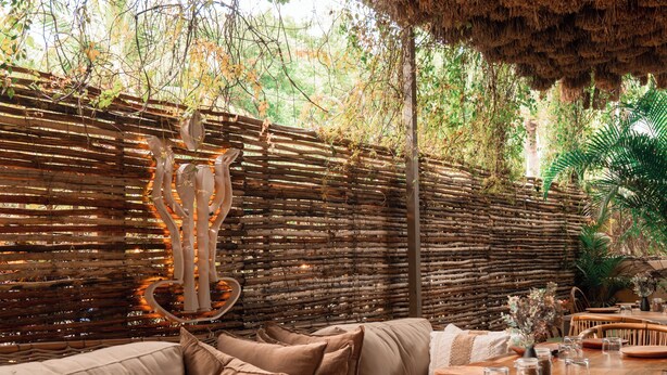 Un espace de restauration en plein air avec des tables et des chaises en bois, un mur en osier et une sculpture décorative.