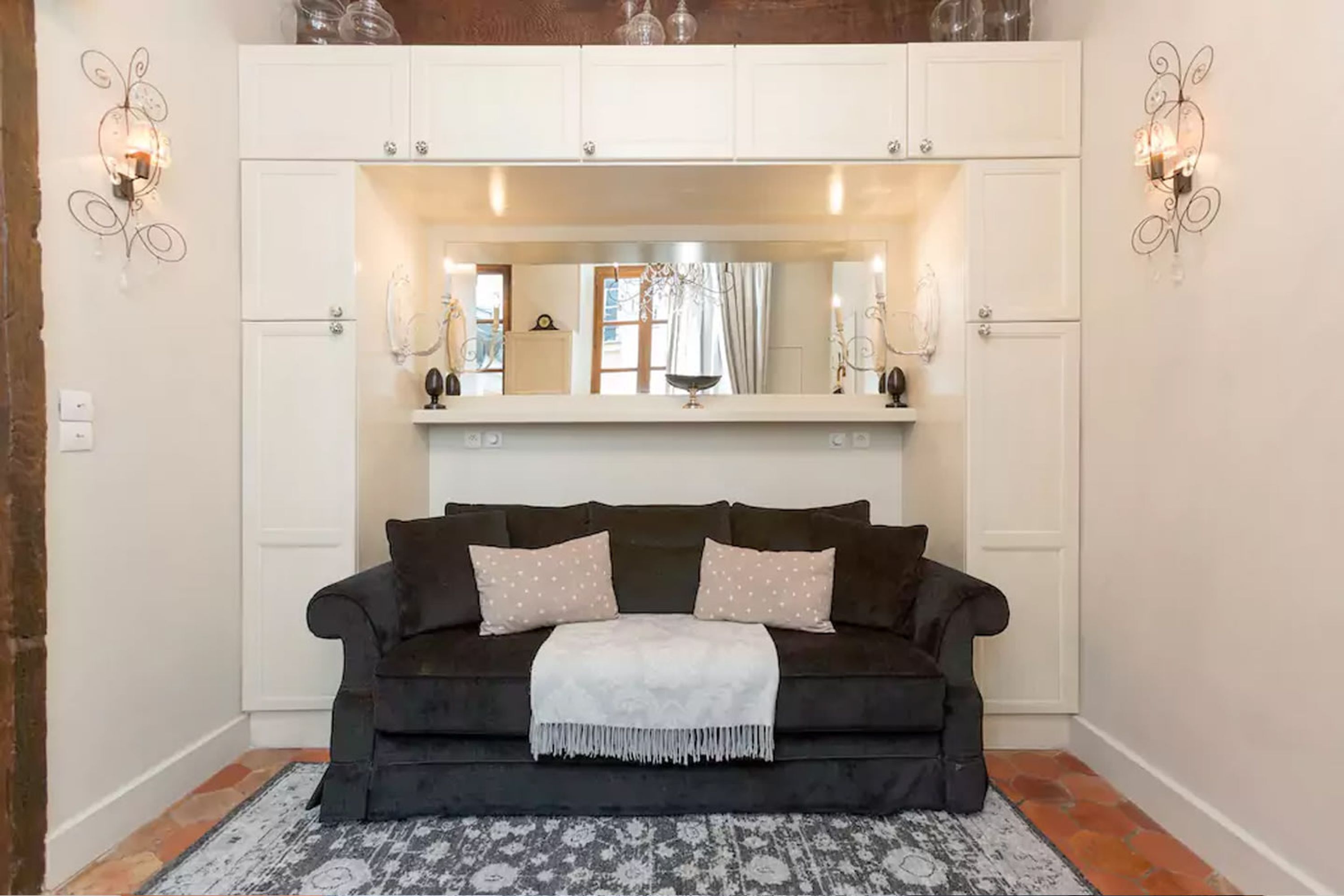 A dark brown sofa with a white throw blanket, a patterned rug, and a mirror above a white cabinet with shelves.