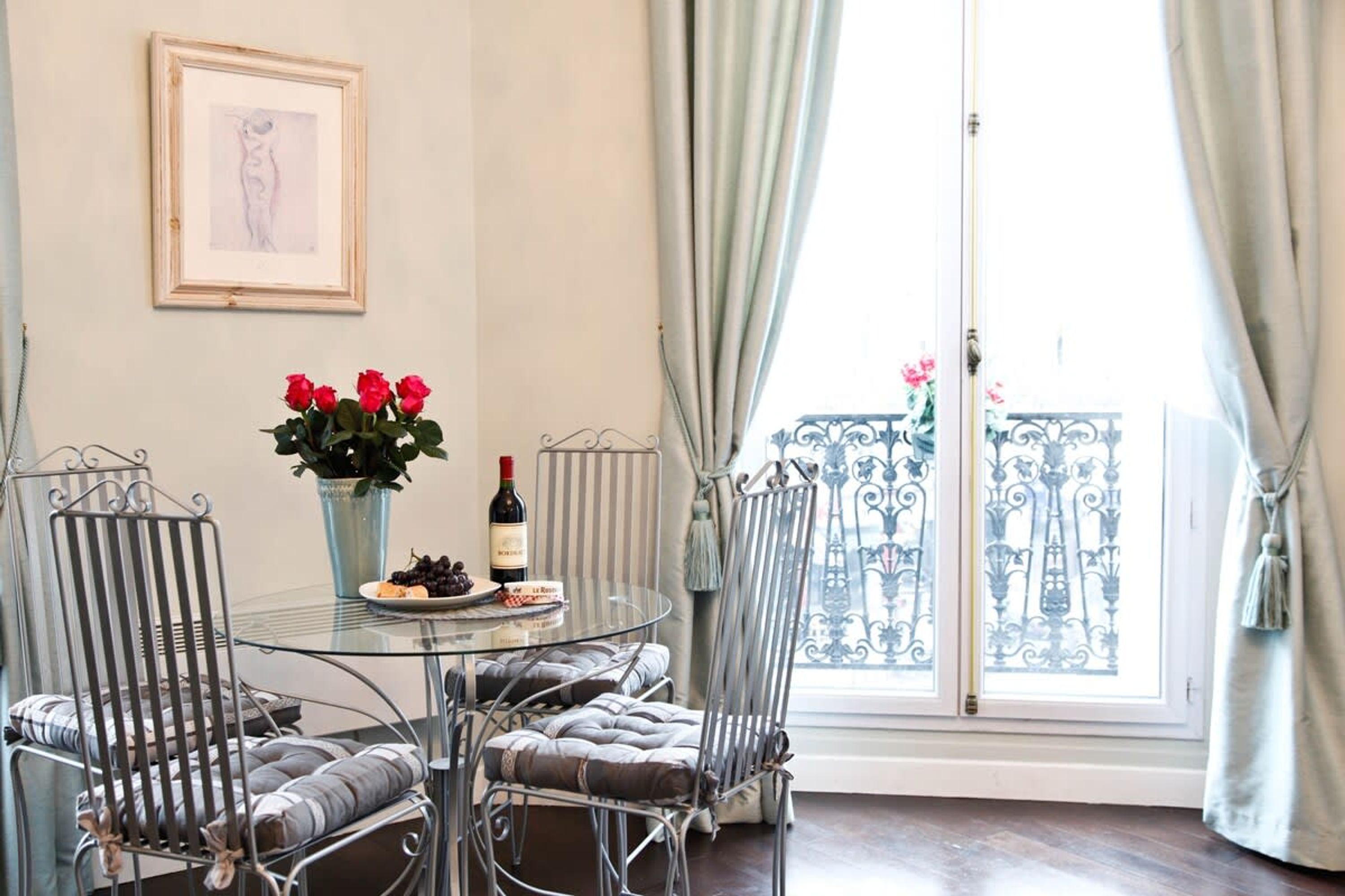 A dining area with a glass table, metal chairs, a vase of red flowers, a bottle of wine, and a framed picture on the wall.