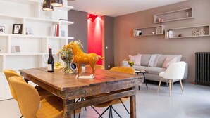 A modern living room with a wooden dining table, yellow chairs, a red accent wall, and a sofa with a white cushion. - Le Gourmet du Marais (Paris)