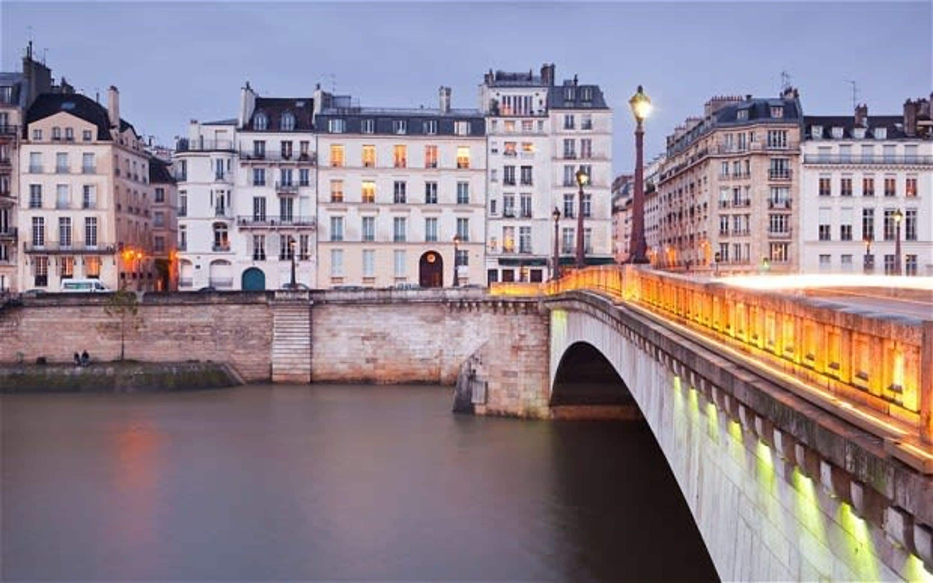 A stone bridge over a calm river, connecting two sides of a city with historic buildings.