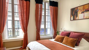 A bedroom with a large bed, orange curtains, a window with blinds, and a painting on the wall. - La Lumiere Eiffel Walk to Tower (Paris)