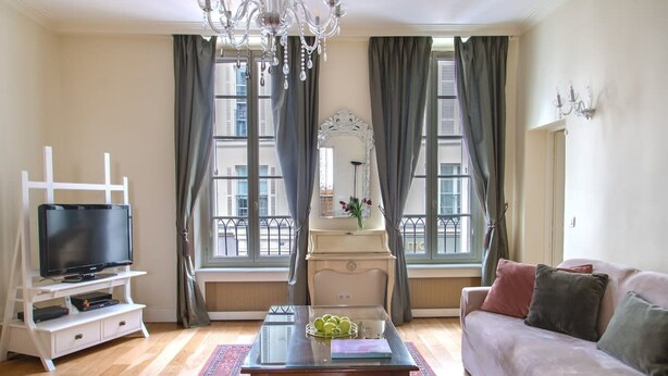 A living room with a television on a white stand, a coffee table with decorative items, a sofa with cushions, and a large window with curtains. - La Lumiere Eiffel Walk to Tower (Paris)