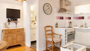 A compact kitchen with white cabinets, a wooden chair, a television, and a clock on the wall.
