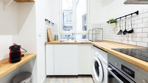 A compact kitchen with white cabinets, a washing machine, a stove, and a window with a view of buildings outside. - Le Loft Latin - New Apt (Paris)